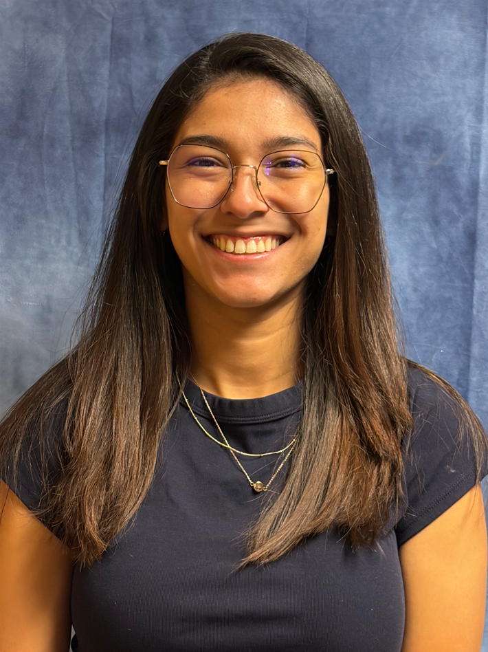 A young woman with long dark hair smiles warmly while wearing glasses and a dark shirt against a blue background.